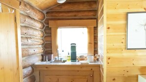 A log cabin kitchen with a wooden interior, a window above the counter, and a power outlet on the wall. - Hideout Cabin, Rustic Hideout Cabin (Story)