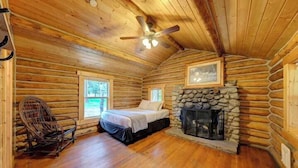A log cabin room with a bed, a rocking chair, a stone fireplace, and a ceiling fan. - Hideout Cabin, Rustic Hideout Cabin (Story)