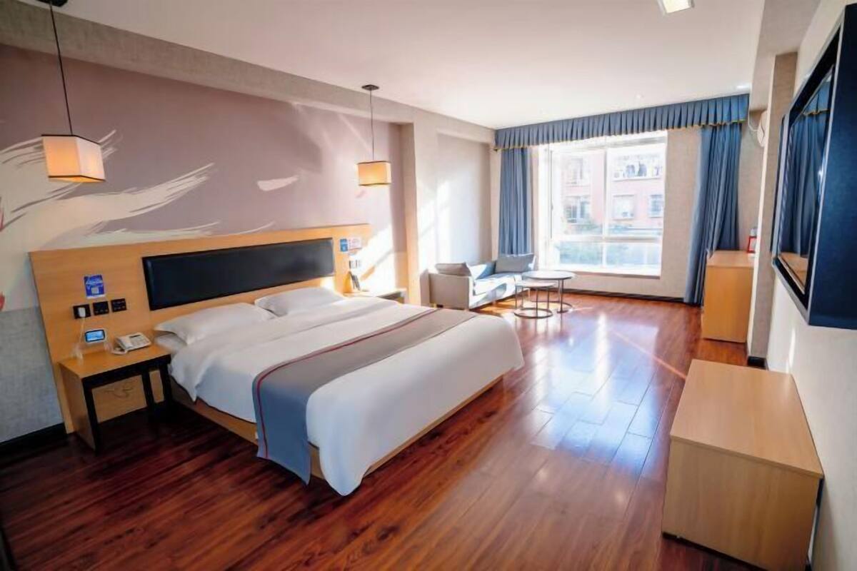 A hotel room with a large bed, bedside tables, a desk, a chair, and a view of buildings through the window.
