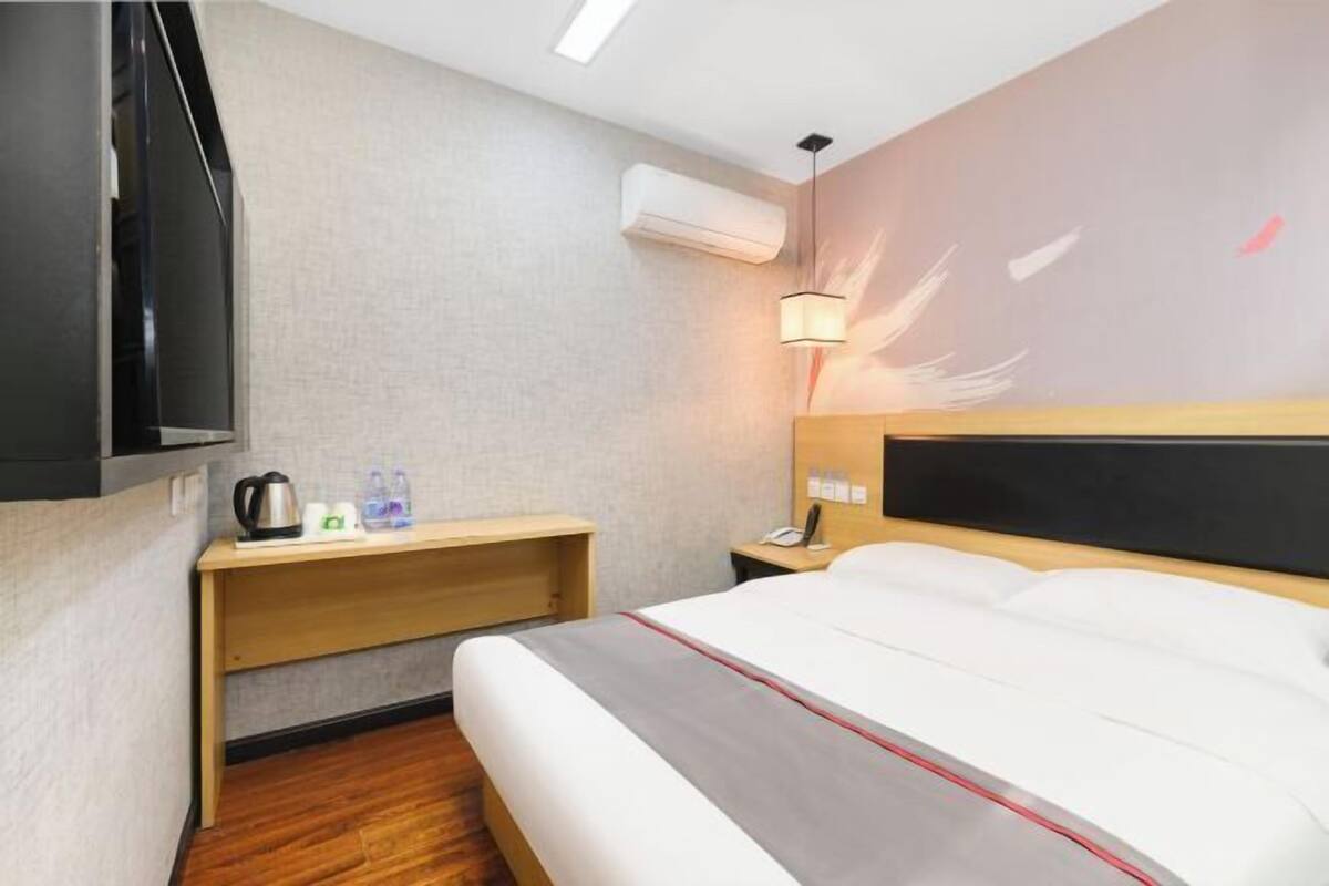 A hotel room with a bed, bedside table, lamp, and an air conditioning unit.