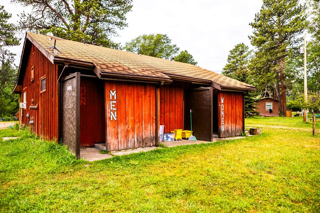Un bâtiment rustique en bois, avec l’inscription « HOMMES » sur le côté, situé dans une zone herbeuse, avec des arbres en arrière-plan.