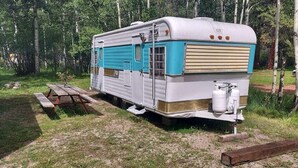 A vintage camper parked in a wooded area with picnic tables nearby. - Mid-Century Holiday Trailer, Rustic Mid-Century RV (Story)