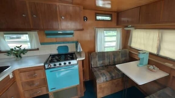A compact RV kitchen with wooden cabinets, a blue stove, and a small dining area with a bench. - Mid-Century Holiday Trailer, Rustic Mid-Century RV (Story)