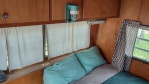 A compact RV interior with a bed, curtains, a window, and a kitchenette area. - Mid-Century Holiday Trailer, Rustic Mid-Century RV (Story)