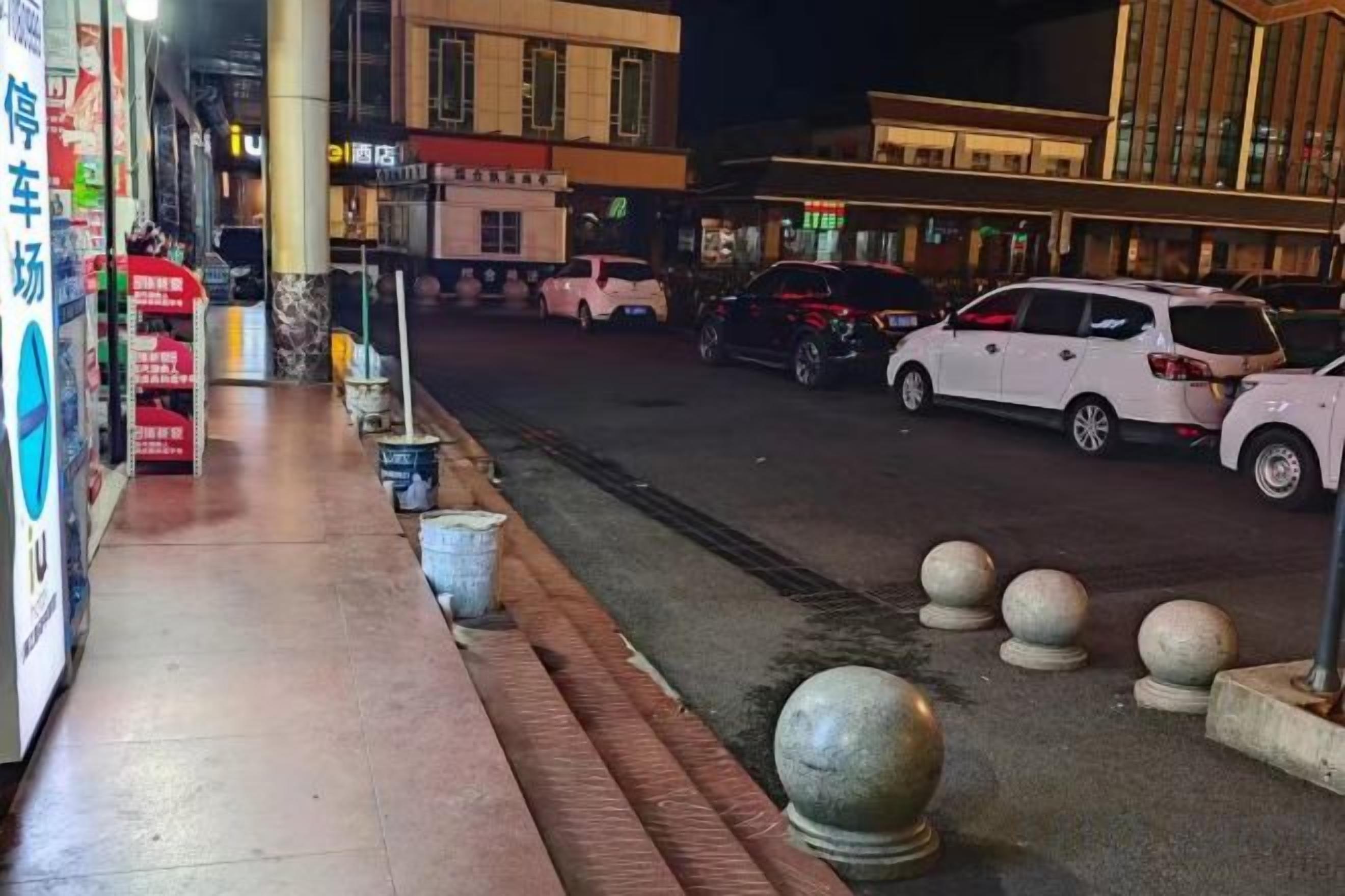 A street view at night with a sidewalk, parked cars, and buildings with illuminated signs.