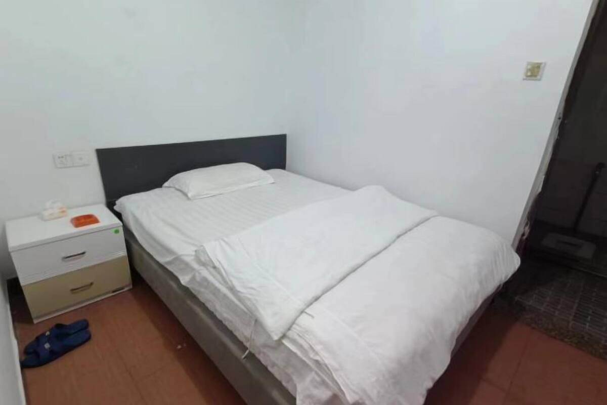 Ein kleines Zimmer mit einem Einzelbett, einer weißen Bettdecke, einem Nachttisch mit Schubladen und einem Paar blauer Hausschuhe auf dem Boden.