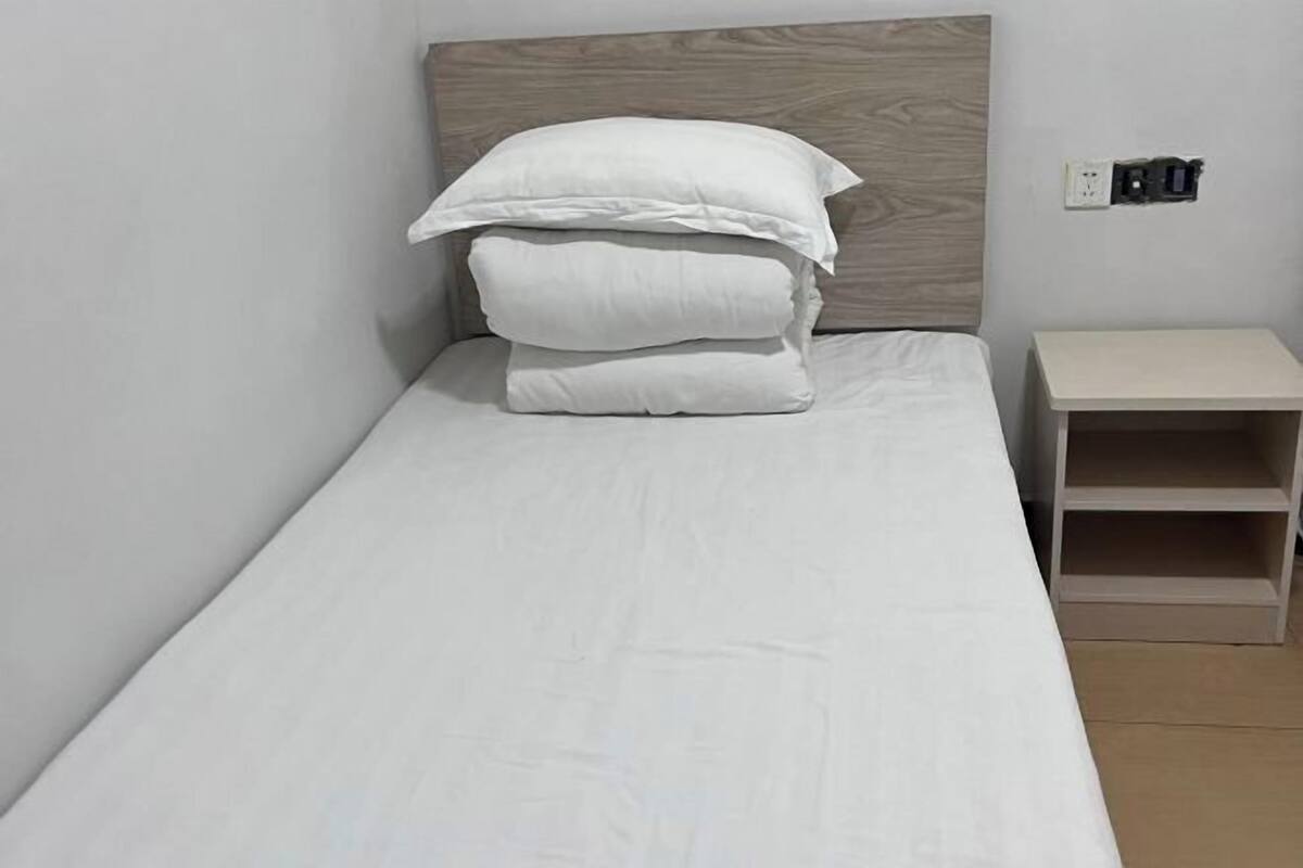 Ein Einzelbett mit weißer Bettwäsche und Kissen, ein Holz-Kopfstück, ein Nachttisch mit Ablage und eine Steckdose.