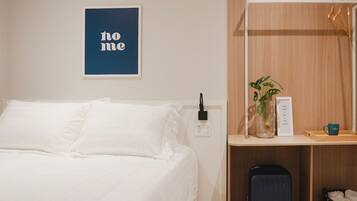 A neatly made bed with white linens, a small bedside table with a plant and a mug, a suitcase on the floor, and a wall-mounted sign with the text "no me".