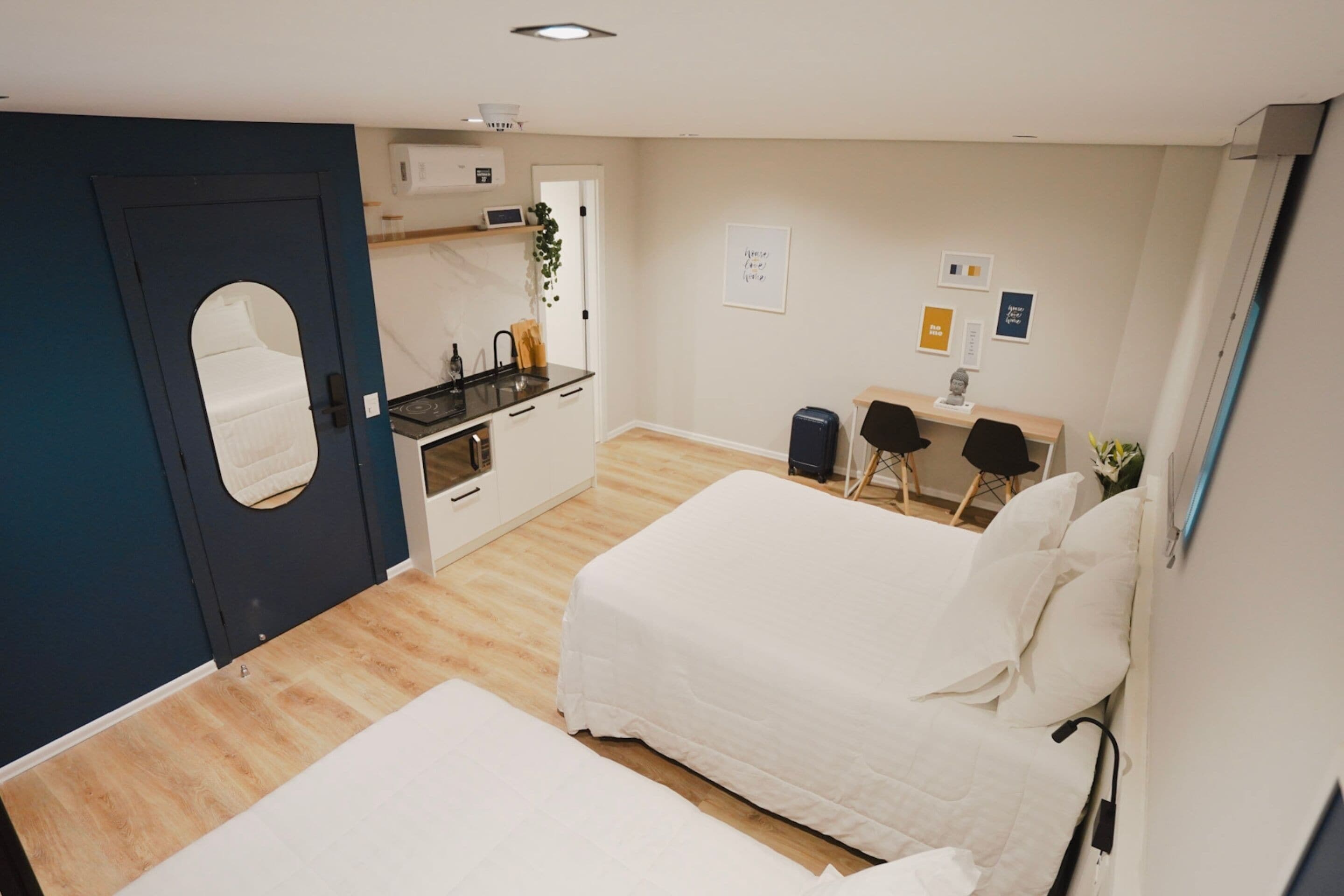 A modern hotel room with a large bed, a smaller bed, a kitchenette, a mirror, and a desk.