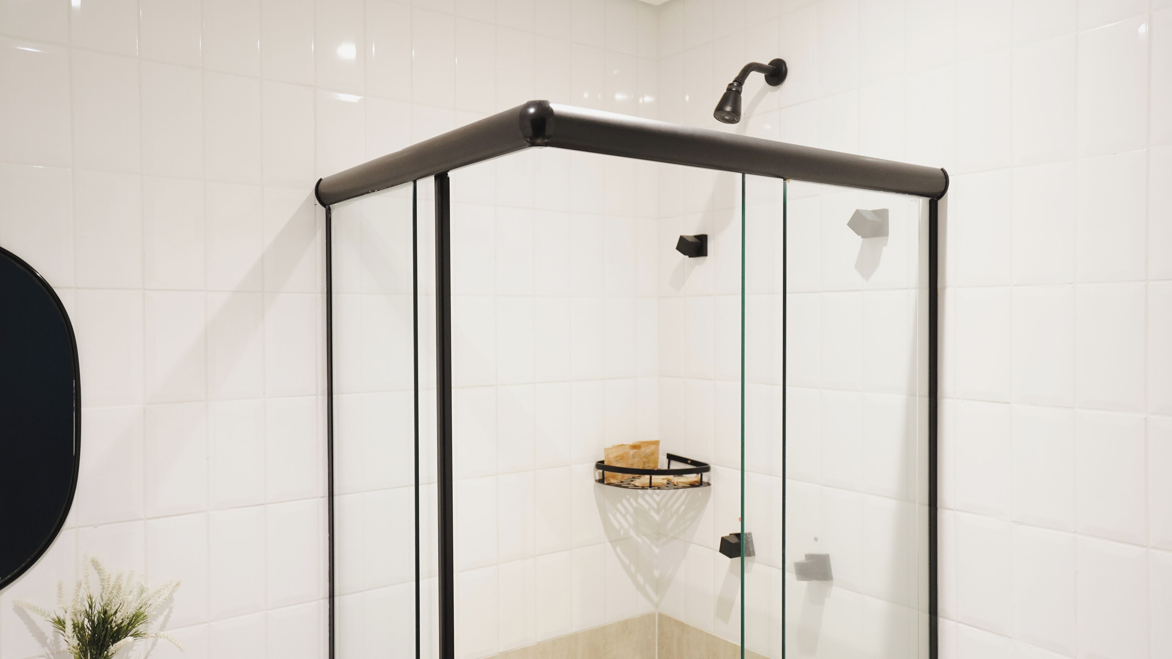 A glass-enclosed shower with a black frame, white tiled walls, and a small shelf with toiletries.