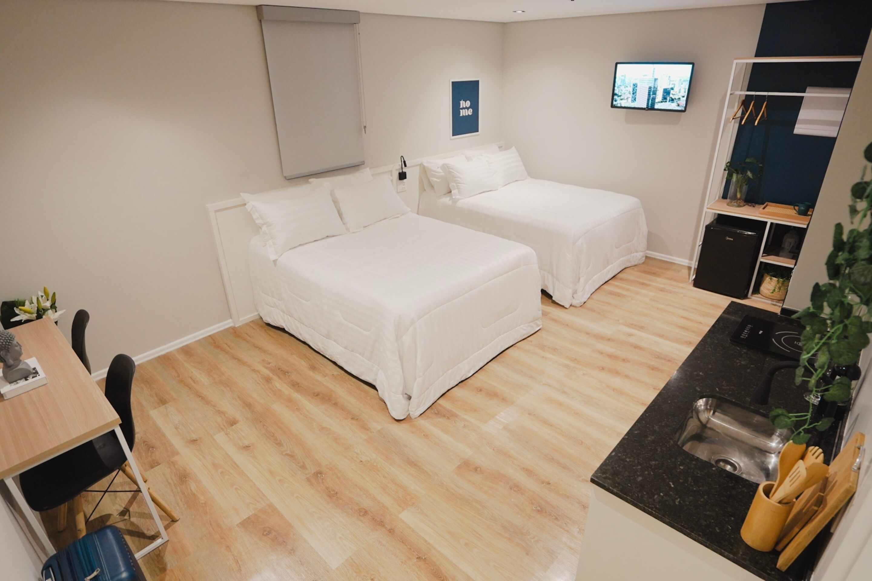 A hotel room with two beds, a desk, a chair, a small kitchenette, and a TV on the wall.