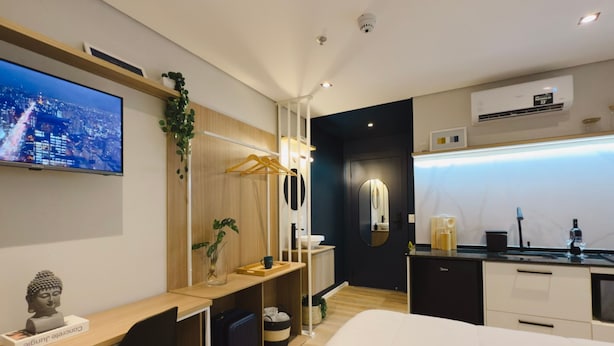 Ein modernes Hotelzimmer mit Bett, Schreibtisch und Sitzecke. Ein Fernseher ist an der Wand angebracht, eine kleine Buddha-Statue steht dort, ebenso eine Pflanze. Das Zimmer verfügt über eine dunkle Akzentwand und ein Fenster mit Blick auf die Stadt.