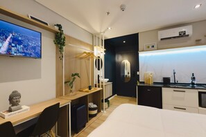 A modern hotel room with a bed, desk, and seating area. There is a TV mounted on the wall, a small Buddha statue, and a plant. The room has a dark accent wall and a window with a view of the city. - Apê Brás - Shopping Plaza Polo (São Paulo)