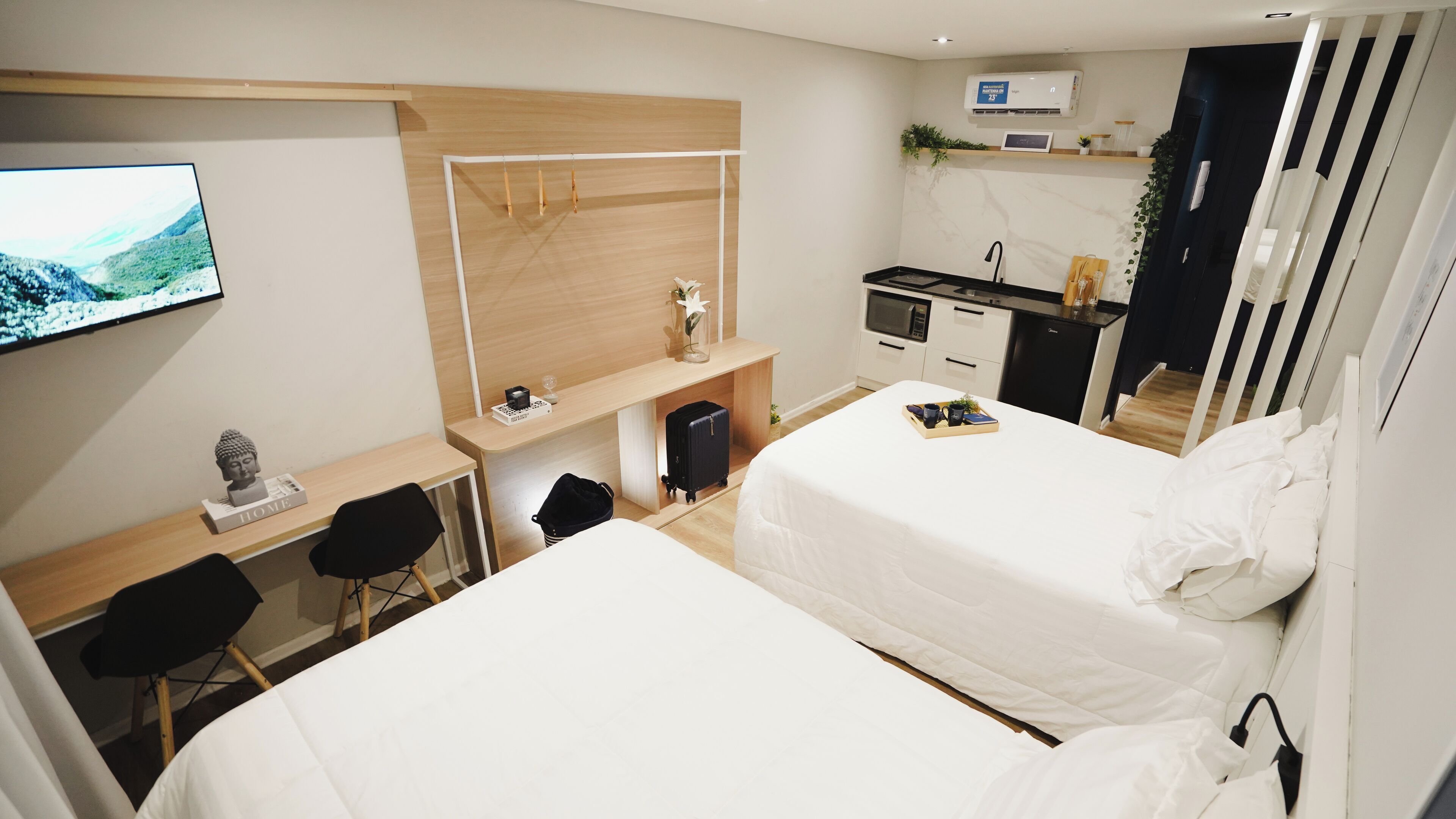 A hotel room with two beds, a desk, a chair, a TV, a kitchenette, and a small bathroom.
