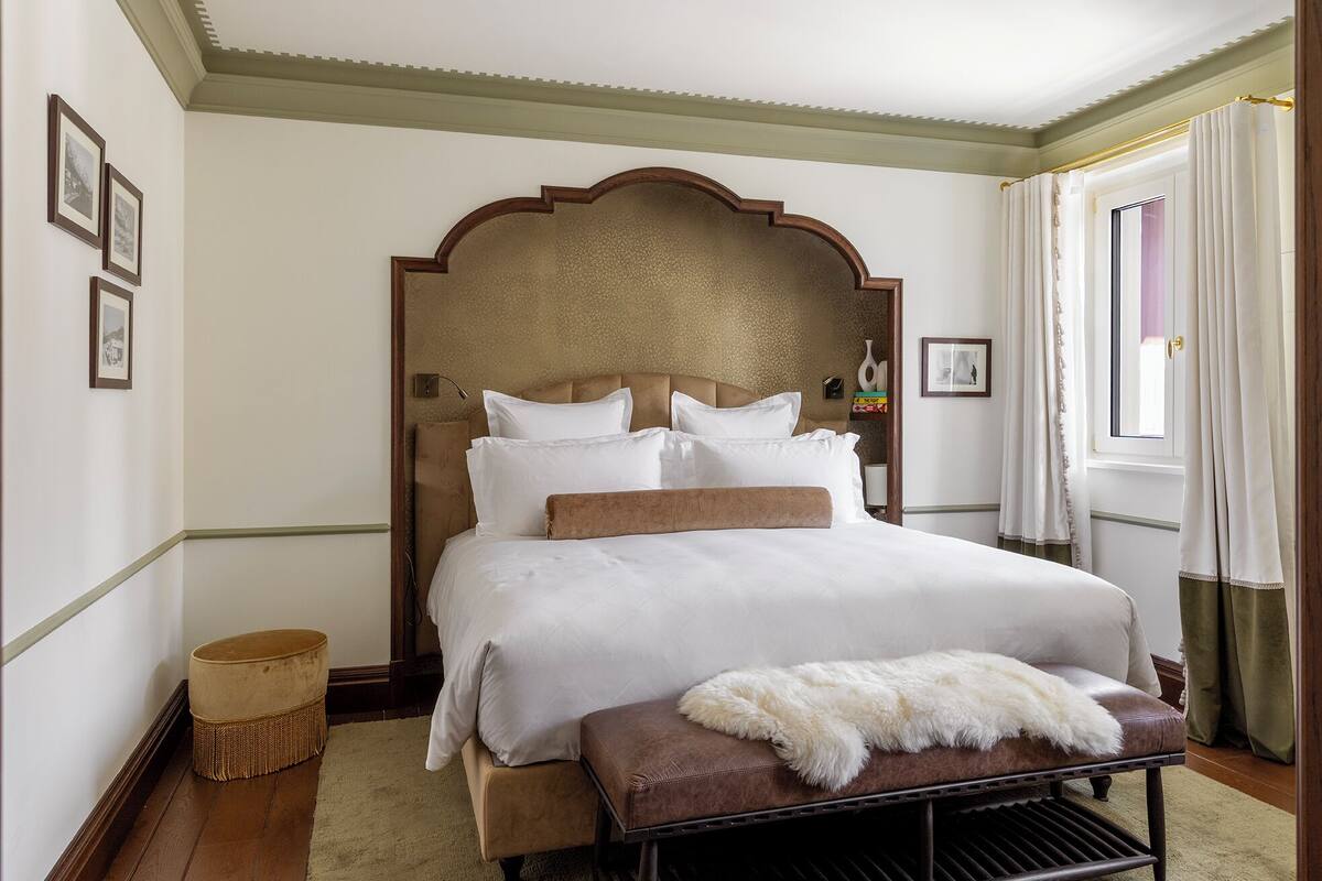 A neatly arranged bedroom with a large bed, a wooden headboard, a small bench, and a window with curtains.