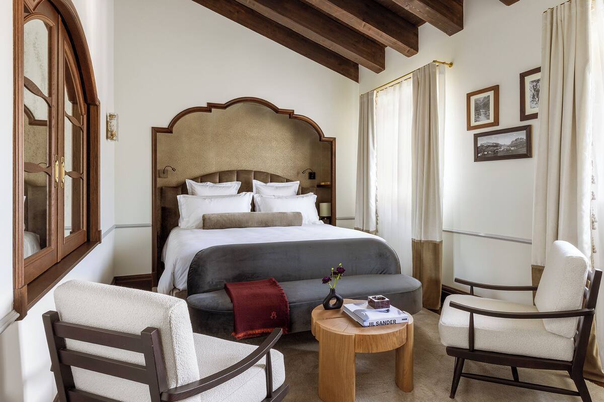 A hotel room with a large bed, two armchairs, a small round table, and a wooden ceiling.
