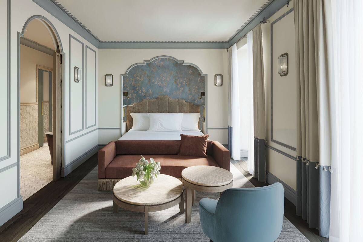 A hotel room with a large bed, a sofa, a small round table, and a chair. There is a door leading to another room and a window with curtains.