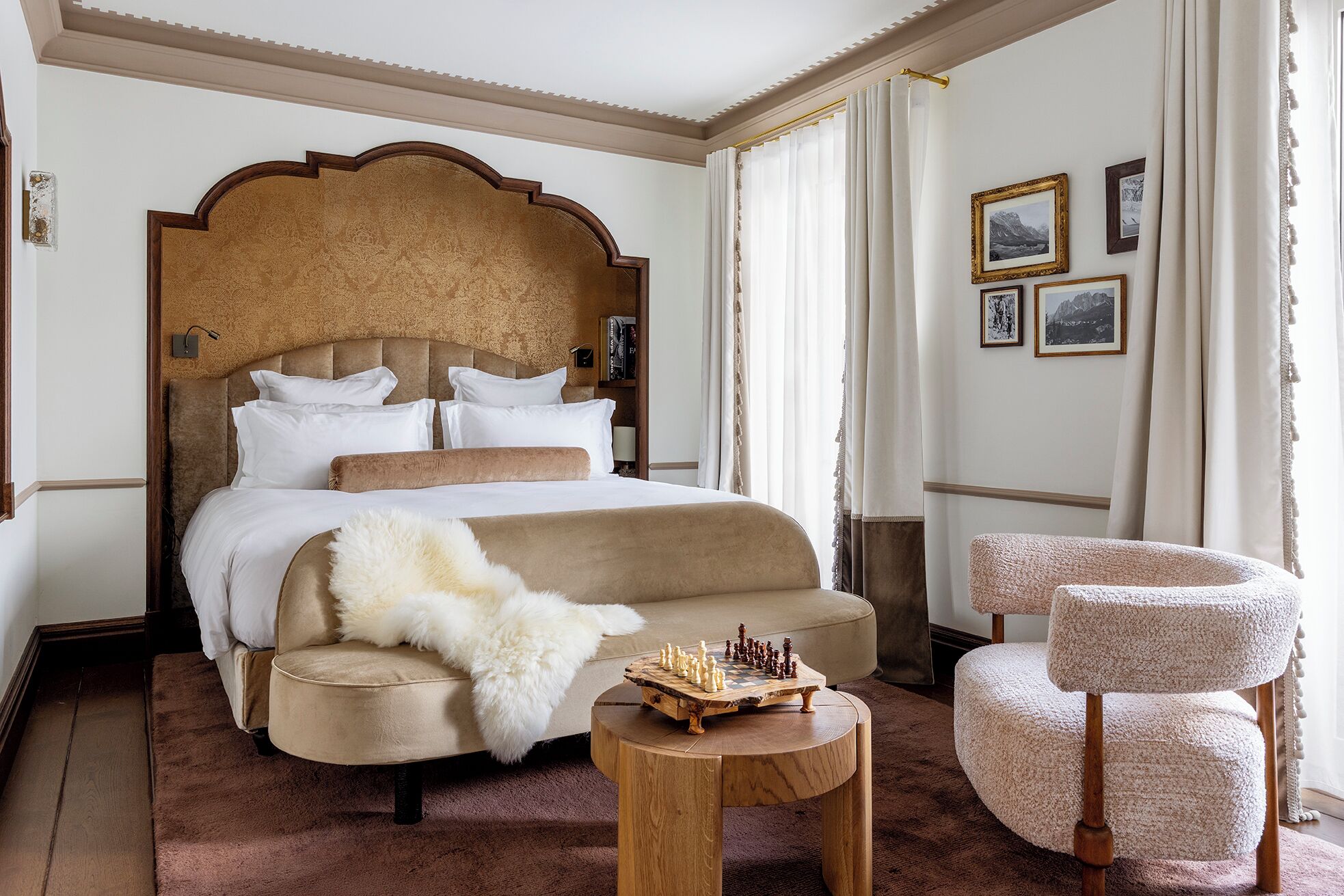 A hotel room with a large bed, a round wooden side table, a plush armchair, and a bookshelf.