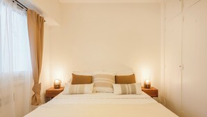 A neatly made bed with a quilted cover, two bedside lamps, and a window with sheer curtains.