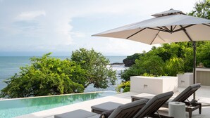 Una piscina infinita con sillas de descanso y una sombrilla, con vistas a un litoral rocoso y abundante vegetación.