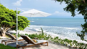 Un salón junto a la playa con una sombrilla blanca, sillones de madera y vistas al océano.
