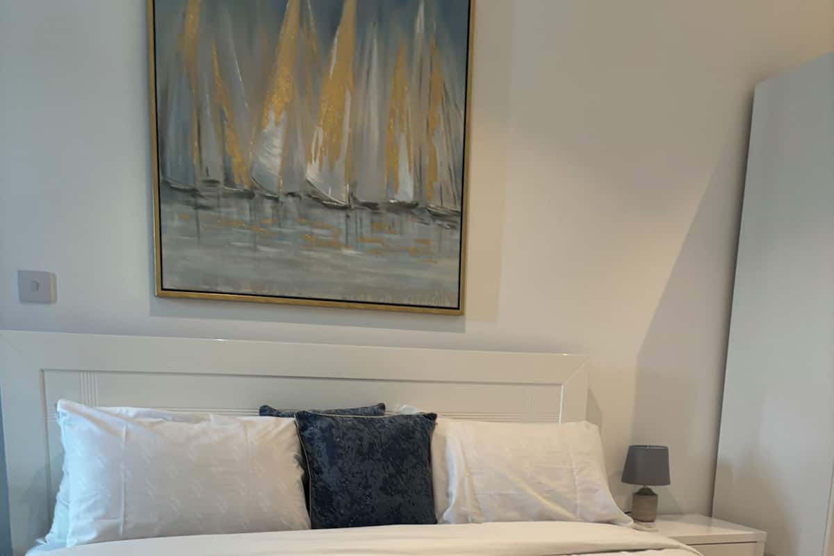 Un lit bien fait, avec du linge de lit blanc et une serviette pliée, une table de chevet avec une lampe et un tableau représentant des voiliers accroché au mur.