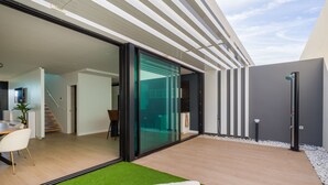 A modern outdoor living space with a glass door leading to a kitchen, a wooden deck, and a small garden area with artificial grass and two white lounge chairs. - Sunset Beach Chalet in Pozo Izquierdo Gran Canaria (Santa Lucia)