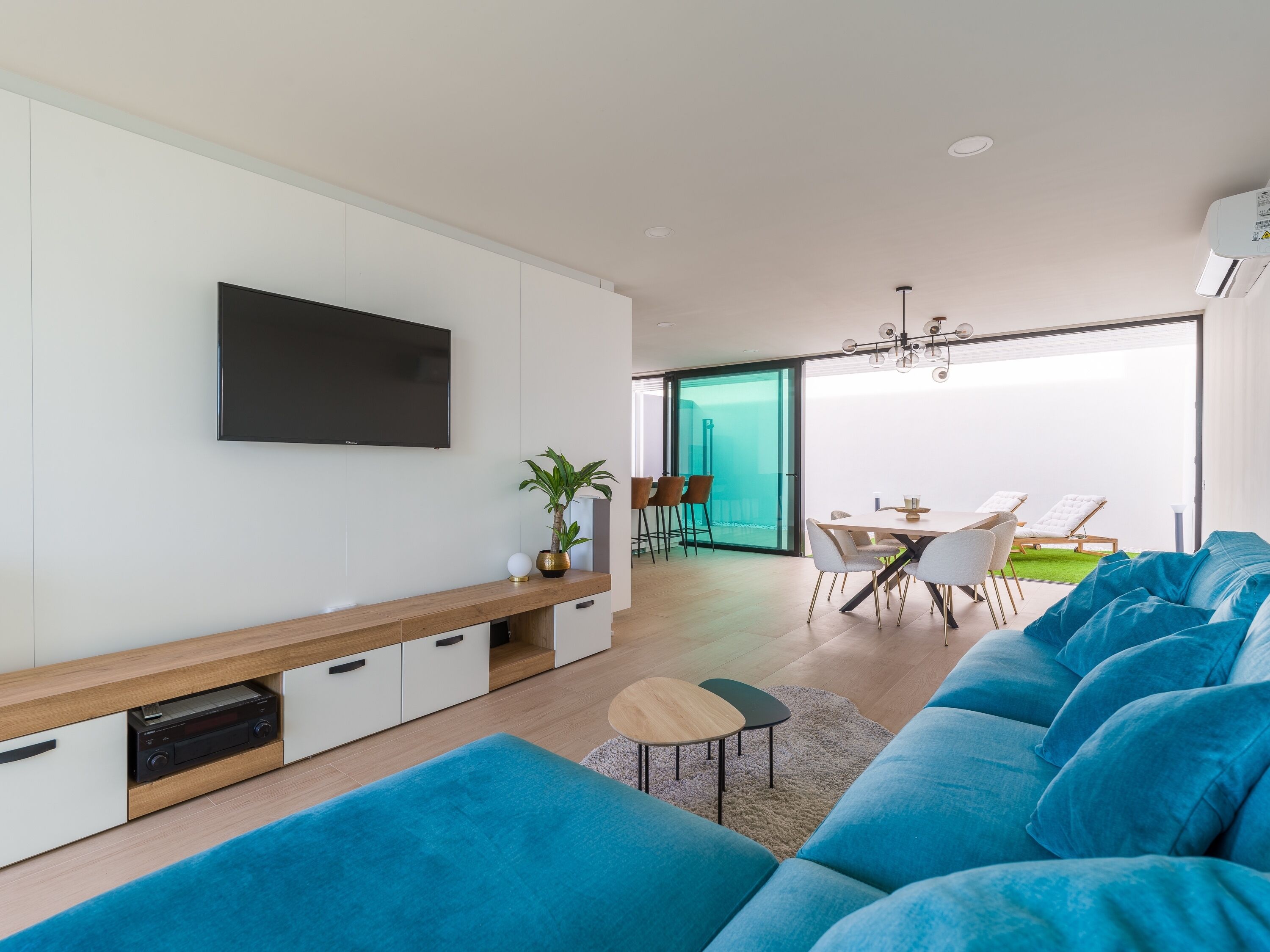 A modern living room with a flat-screen TV, a wooden console table, a blue sofa, a dining area with a glass door leading outside, and a ceiling fan.