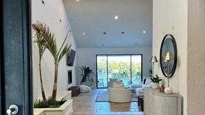 Uma sala de estar ampla com um sofá branco curvo, uma mesa de apoio com objetos decorativos e uma grande janela com vista para o verde.