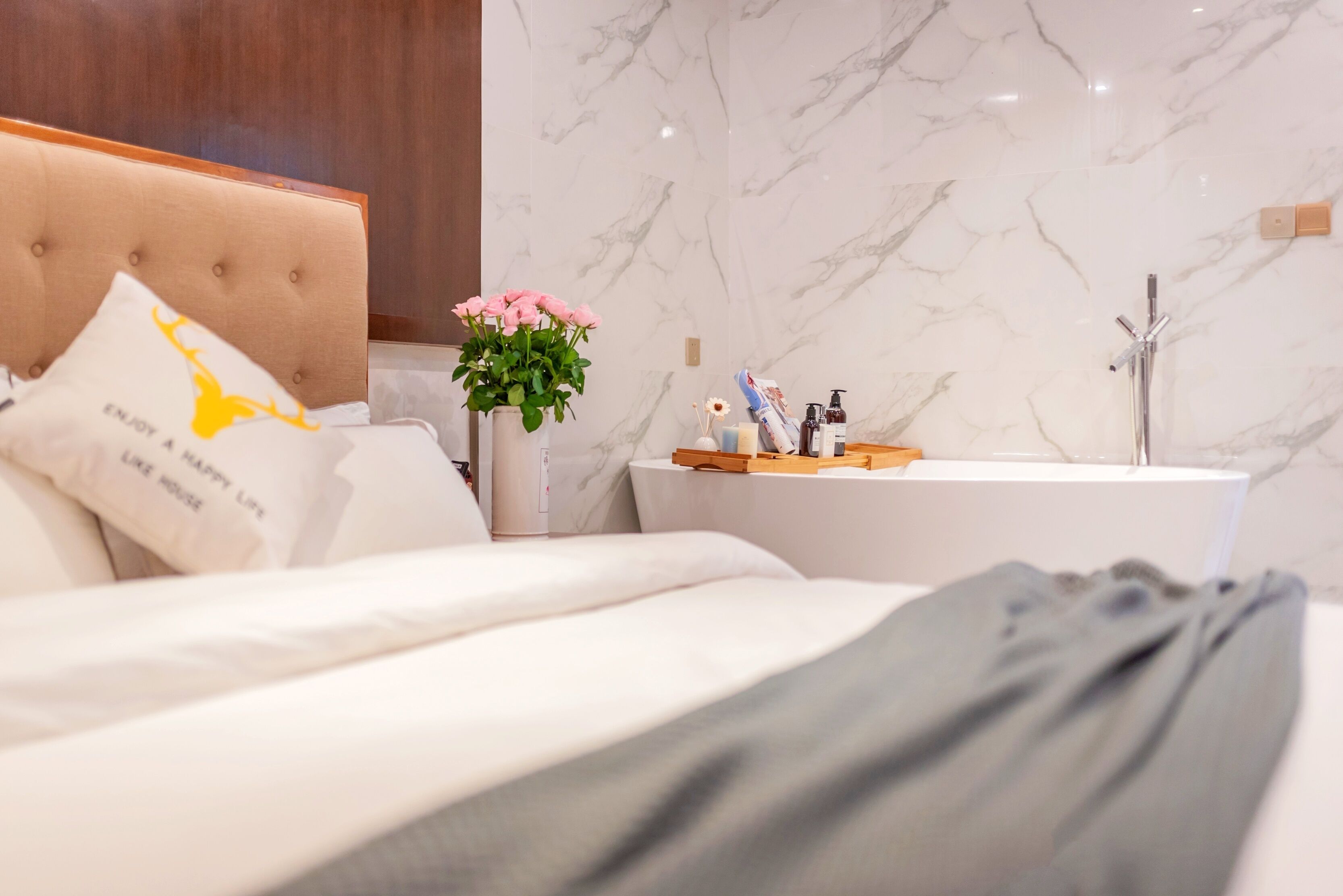 A modern hotel room with a tufted headboard, a bed with white bedding, a bedside table with a vase of pink flowers, and a bathroom with a marble wall and a bathtub.