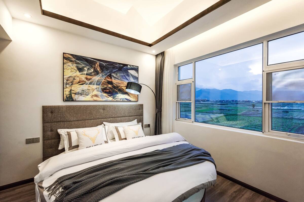 A modern bedroom with a large bed, a cityscape view through a window, and a contemporary abstract painting on the wall.