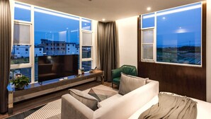 A modern living room with a large flat-screen TV, a sofa, a green chair, and a view of buildings through large windows. - The Field Inn (Dali)