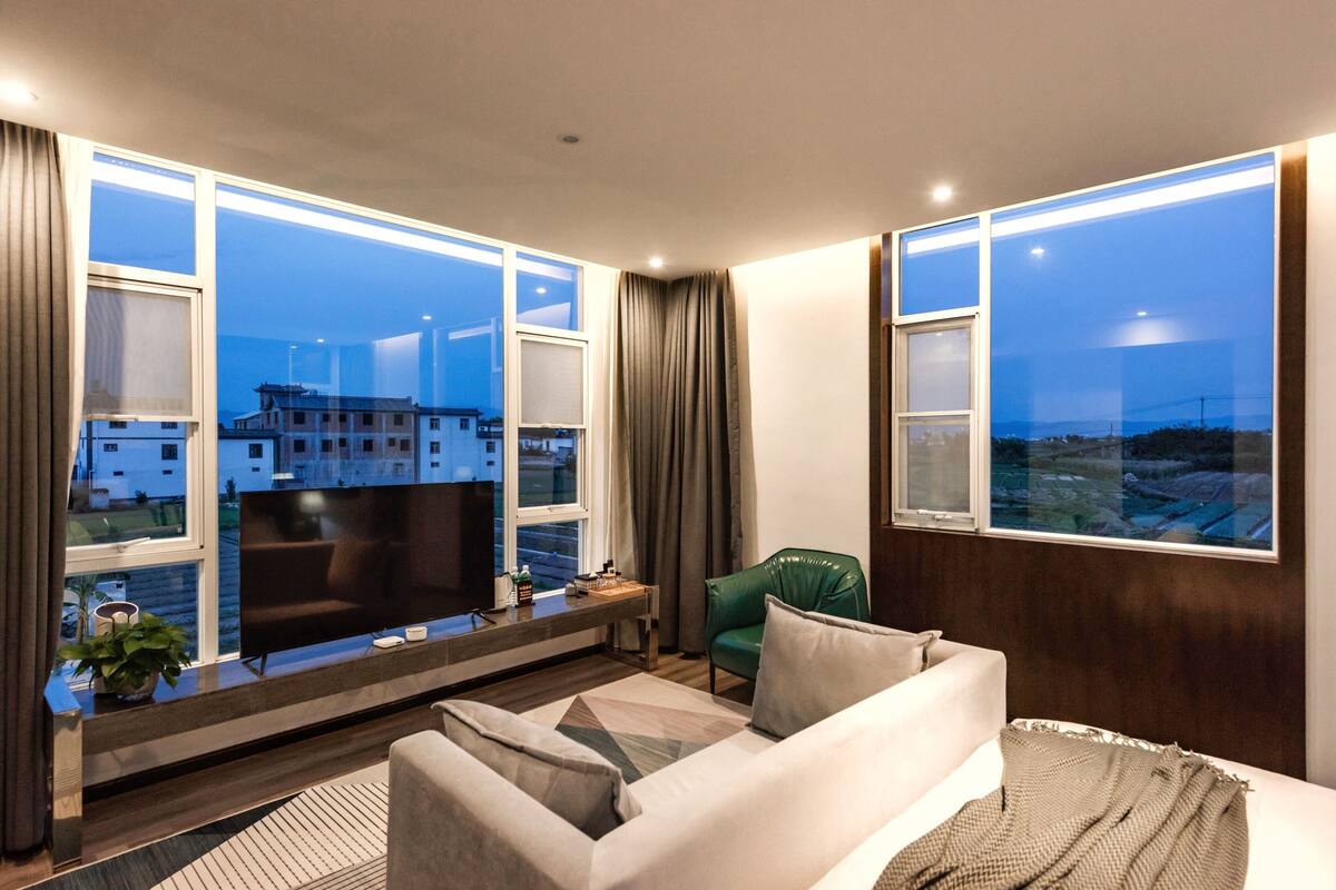 A modern living room with a large flat-screen TV, a sofa, a green chair, and a view of buildings through large windows.