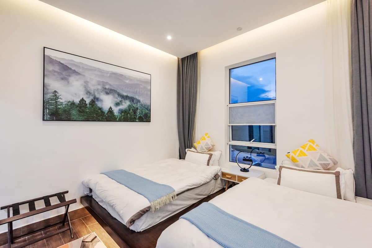 A hotel room with two beds, a desk with a computer, a window with a view of the sky, and a painting of a forest on the wall.