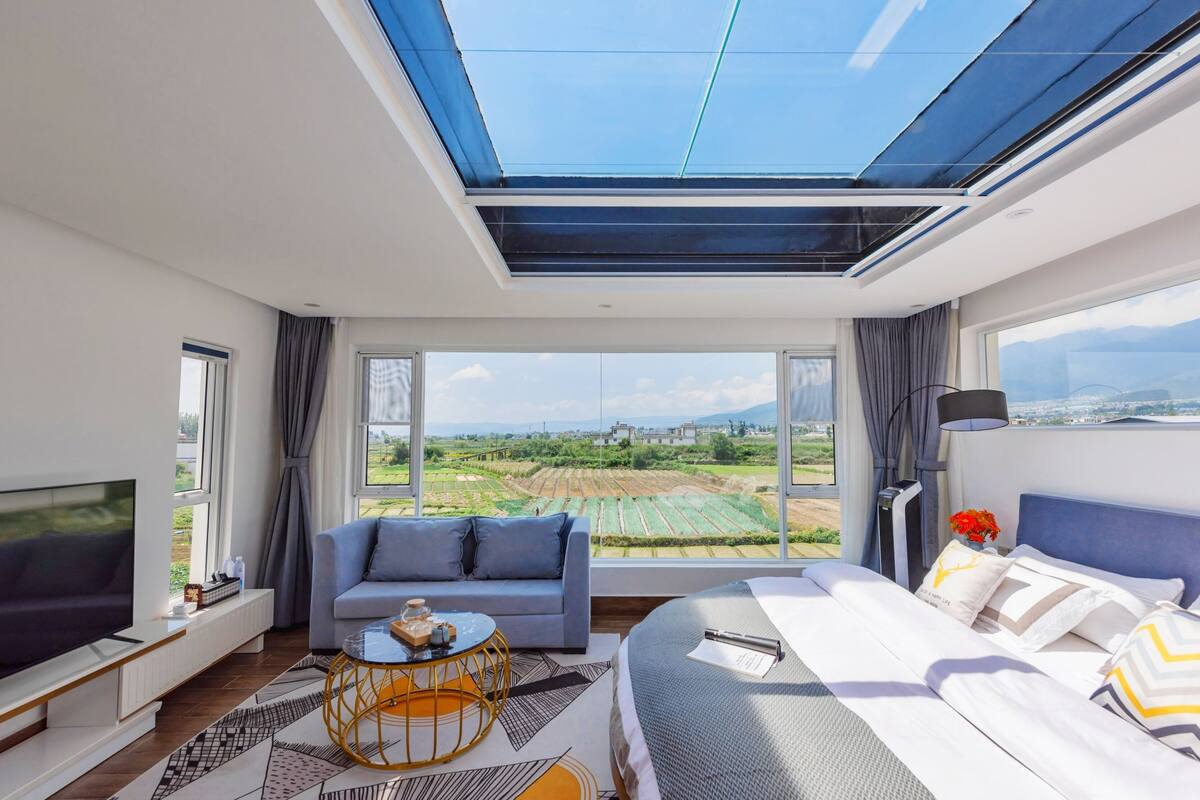 A modern hotel room with a large bed, a sofa, a TV, and a view of the countryside through large windows.