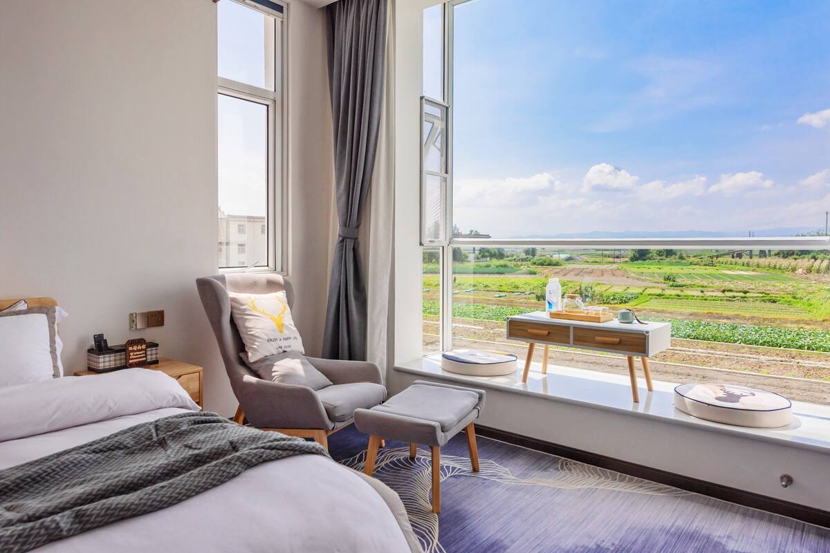 A hotel room with a large window, a bed, a chair, a small table, and a view of a green landscape with a train track.