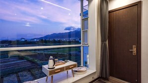 A room with a large window overlooking a cityscape and mountains. There is a small table with a kettle and a cup, and a door with a curtain. - The Field Inn (Dali)