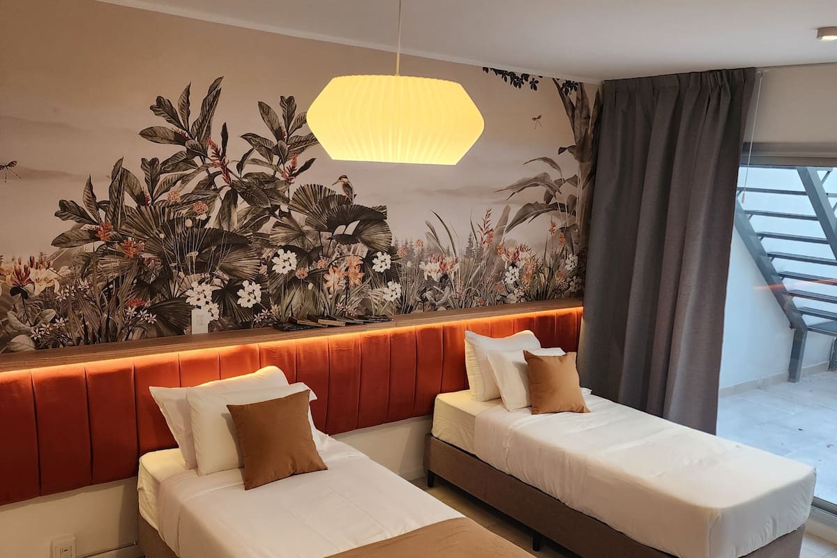 A hotel room with two beds, a large window with curtains, a wall mural, and a hanging light fixture.