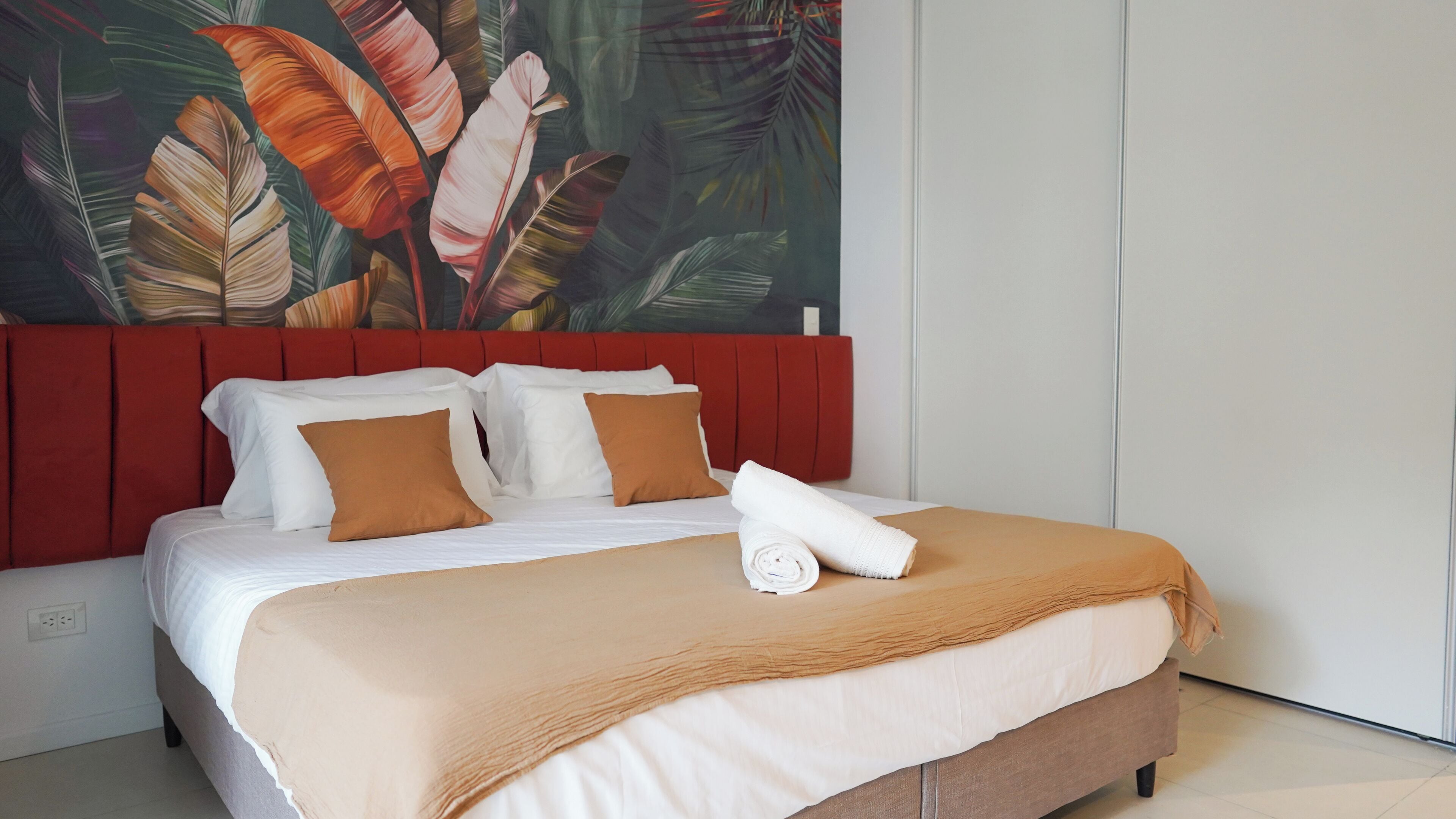 Una cama bien hecha con ropa de cama blanca y marrón, un cabecero rojo y un mural de hojas tropicales en la pared.