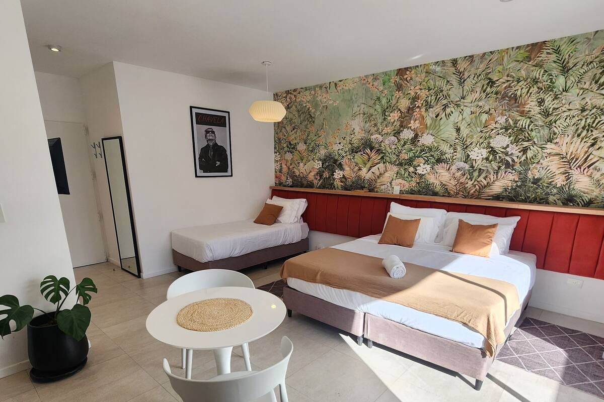 A modern hotel room with two single beds, a dining table, a potted plant, and a large tropical-themed wall mural.