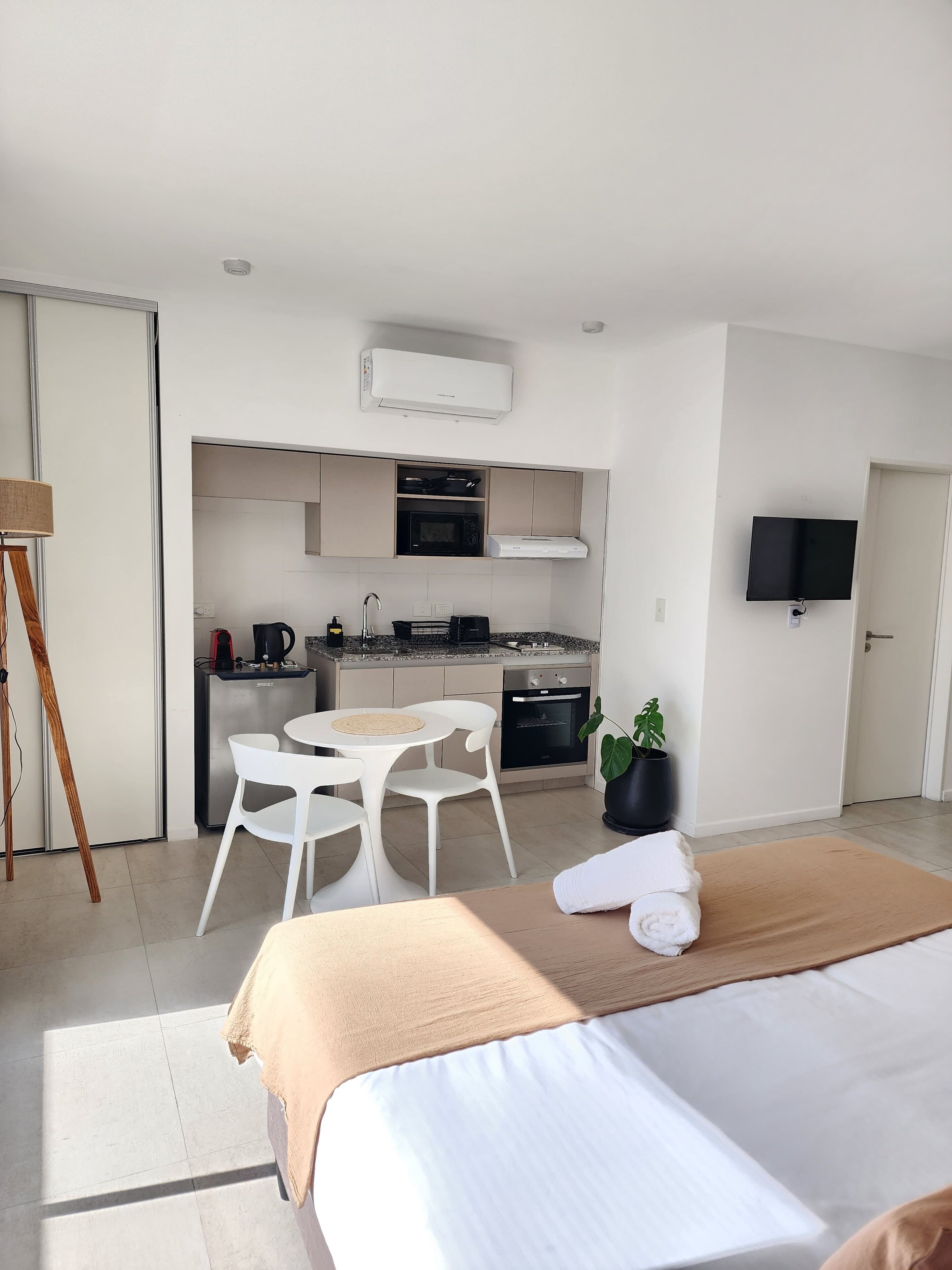 A modern hotel room with a bed, a small kitchenette, a dining area, and a TV.