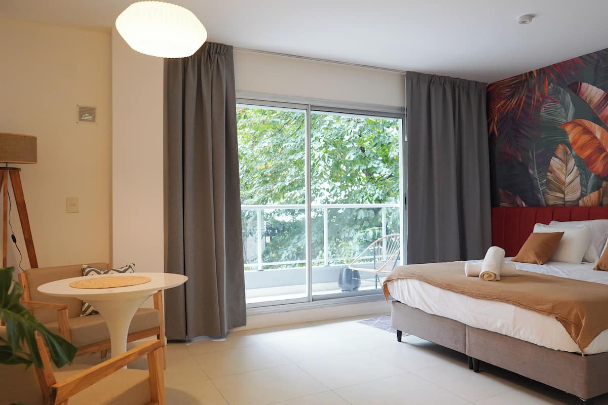 A hotel room with a large bed, a small table, a chair, a large window with curtains, and a tropical leaf mural on the wall.