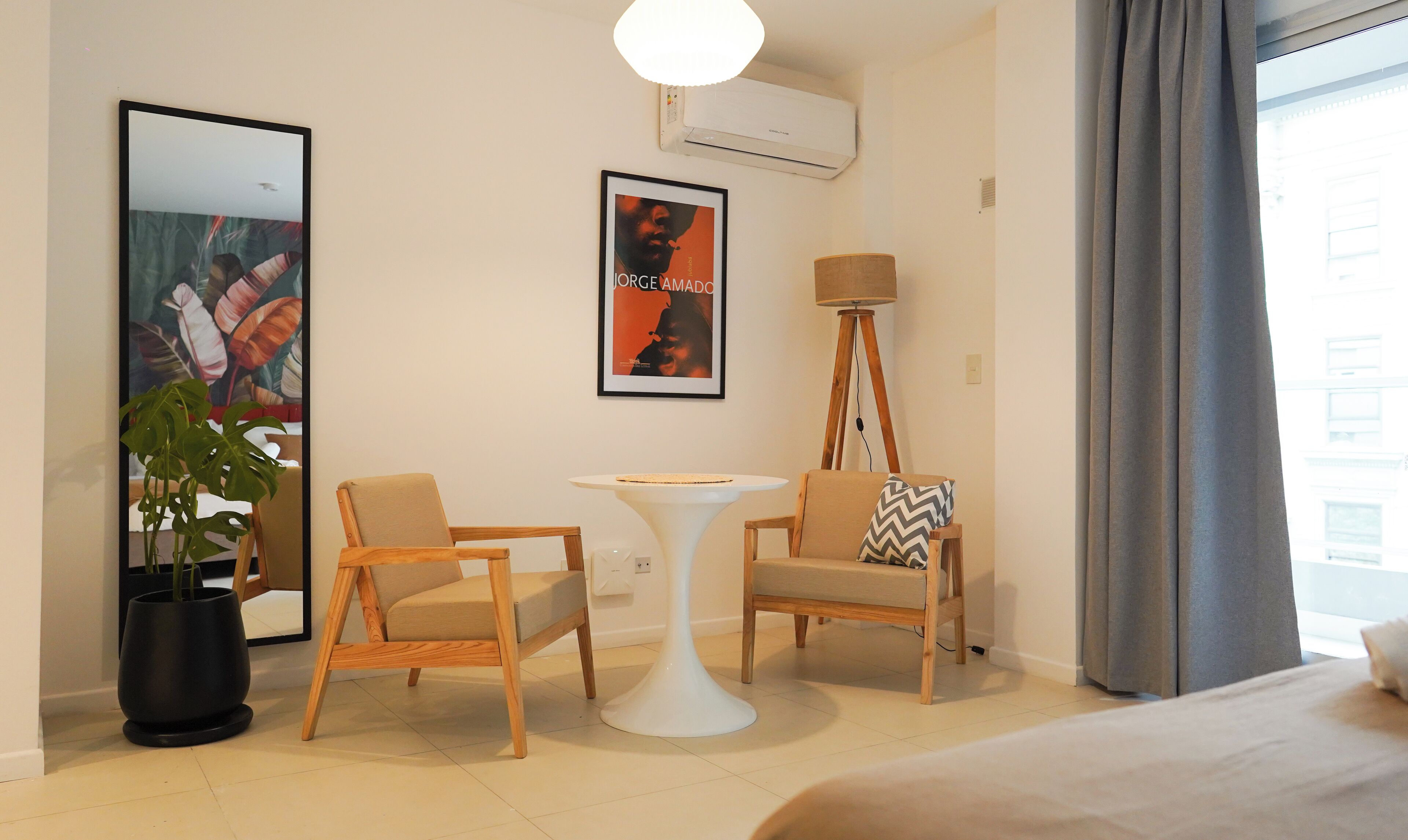 A hotel room with a bed, two wooden chairs, a small white table, a potted plant, a poster, and an air conditioning unit.