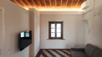 Una stanza con soffitto in legno, una finestra con persiane, una televisione a schermo piatto montata a parete, un pavimento decorato e un divano.