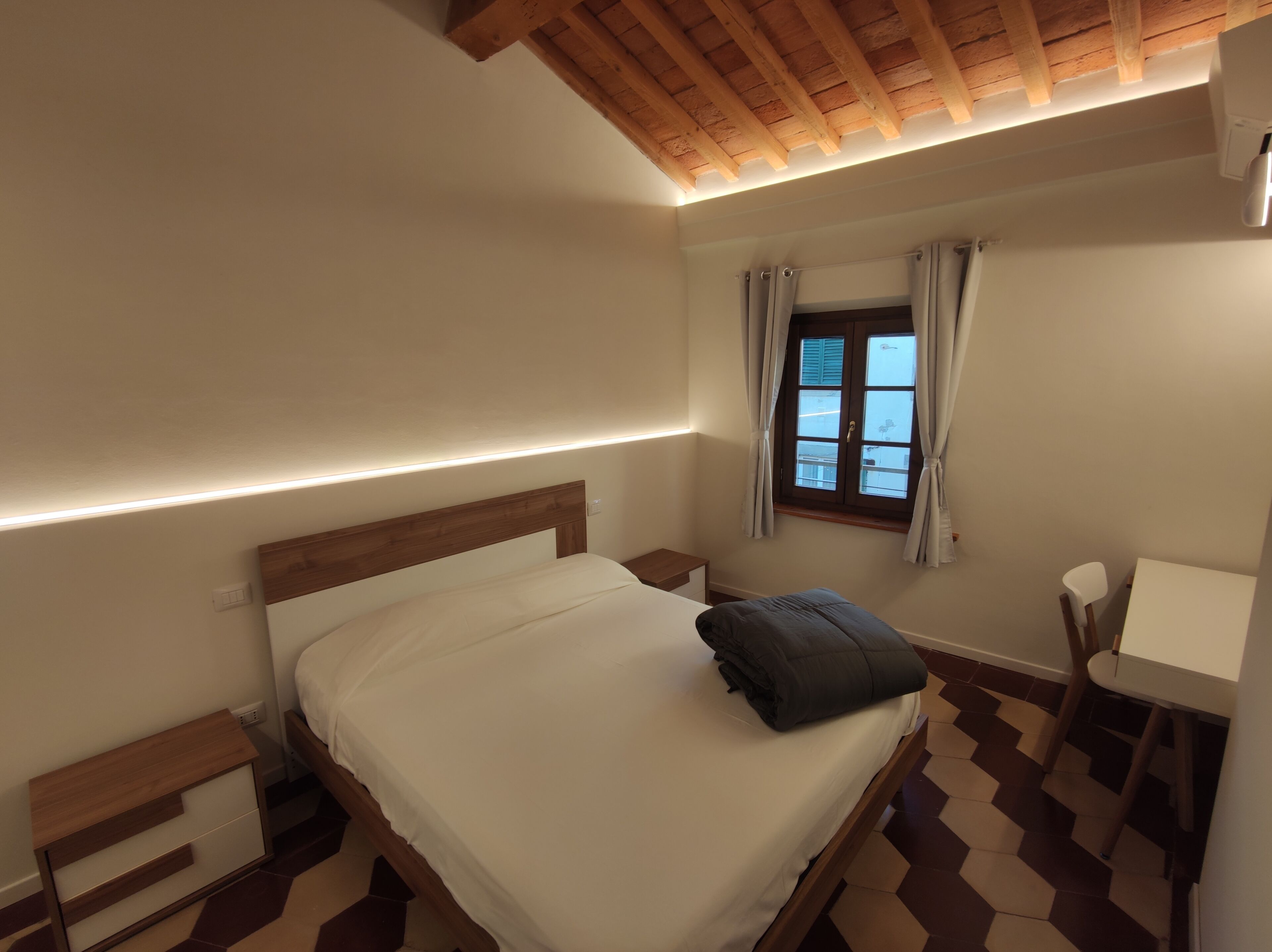 Quarto com cama, mesa de cabeceira e janela com cortinas.