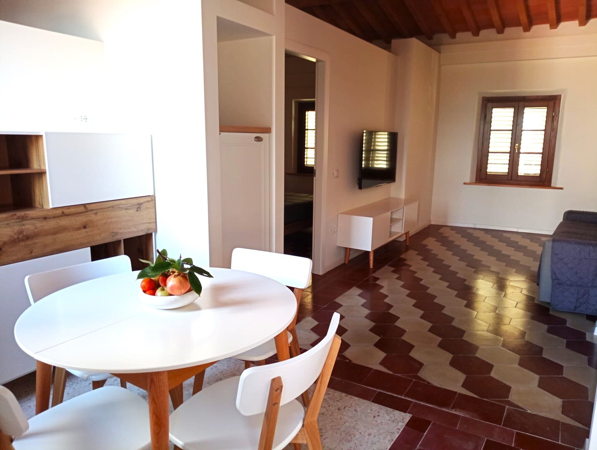 Uma sala de jantar com uma mesa redonda e cadeiras, uma tigela de frutas e uma televisão fixada na parede.