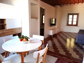 A dining area with a round table and chairs, a fruit bowl, and a television mounted on the wall. - Apartment with a large panoramic terrace in the village center (Peccioli)