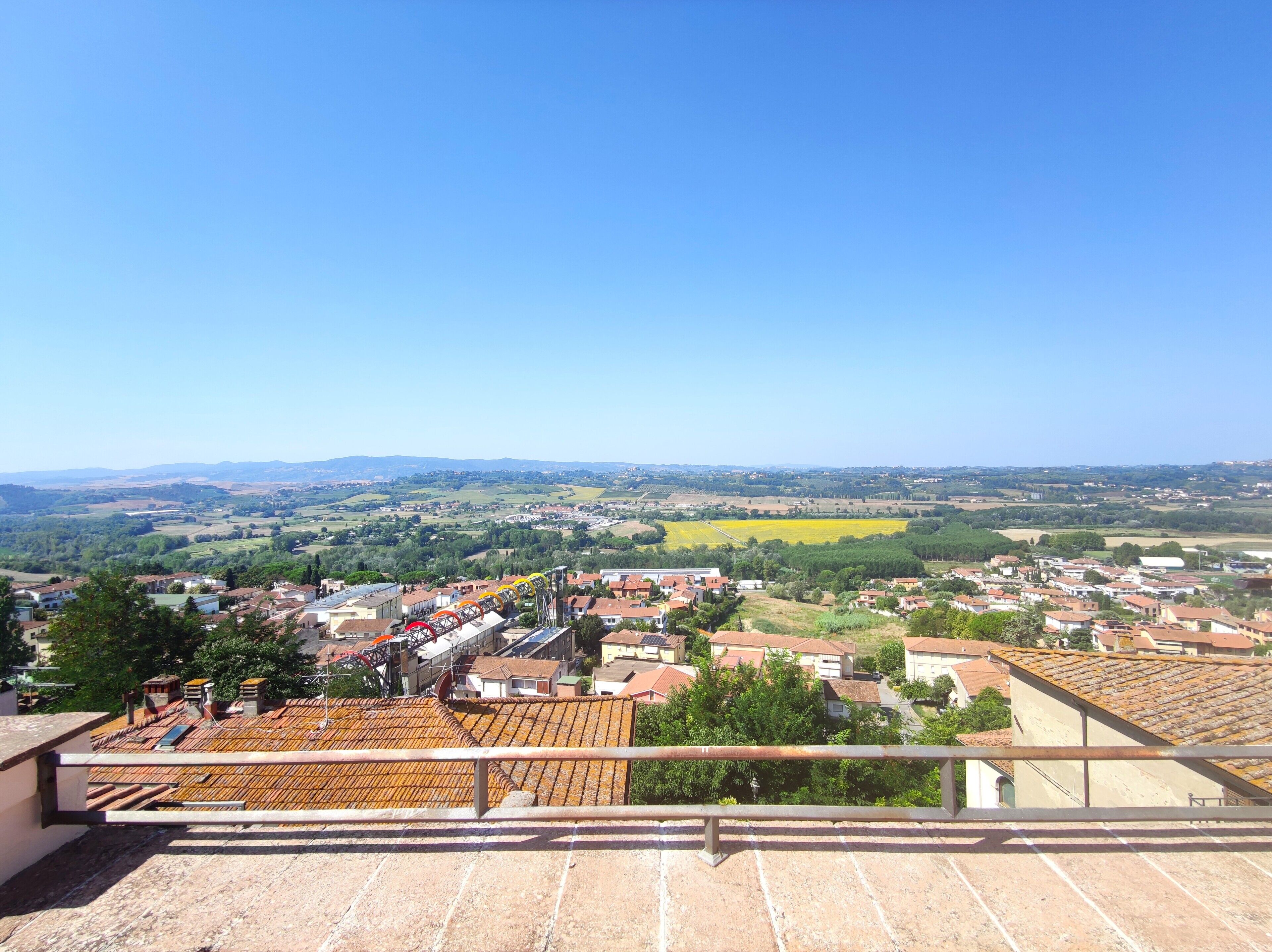 A view from a high vantage point overlooking a town with terracotta-roofed buildings, a clear sky, and expansive fields in the distance.