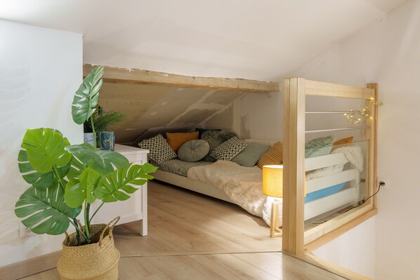 A cozy loft bedroom with a sloped ceiling, a bed with a white and blue bedding, a small white table, and a large green plant in a woven basket.