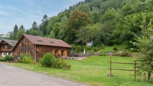 Un bâtiment rustique en bois, doté d’un toit en pente, situé dans une vallée verdoyante, avec un mur de pierres et une clôture en bois.